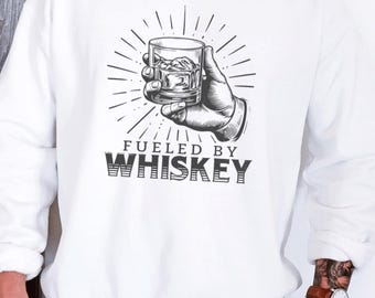 Può includere: Felpa bianca con grafica in bianco e nero di una mano che tiene un bicchiere di whisky con ghiaccio. Il testo "FUELED BY WHISKEY" è stampato sotto. Felpa in stile casual.