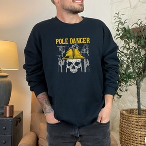 Puede incluir: Una sudadera negra con el texto "POLE DANCER" y una calavera con un casco amarillo. La calavera está rodeada de líneas eléctricas y postes de electricidad.