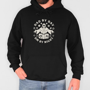 May include: Black hooded sweatshirt with a white graphic of a person in a hooded robe with a book and dice, surrounded by the text "Dad by Day DM by Night".