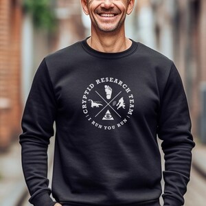 Op de afbeelding: Zwart sweatshirt met een wit grafisch ontwerp van een cirkel met de tekst "Cryptid Research Team I Run You Run" en een gestileerde afbeelding van een voetafdruk, een dinosaurus, een Bigfoot en een oog.