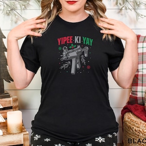 Puede incluir: Camiseta negra de cuello redondo con el texto "YIPEE KI YAY" en rojo y verde, junto con un gráfico de una ametralladora decorada con elementos navideños. Un diseño festivo y humorístico.