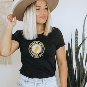 Puede incluir: Camiseta negra con un gráfico amarillo y blanco de un círculo con el texto "Central City Running Club" y un rayo dentro. El texto "Running Faster Than Light Since 1989" está debajo del círculo.