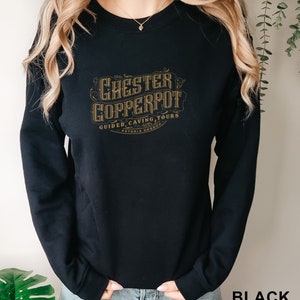 Puede incluir: Sudadera negra con el texto "CHESTER COPPERPOT GUIDED CAVING TOURS ASTORIA OREGON" en un diseño dorado de estilo vintage. La palabra "BLACK" está impresa en blanco en la esquina inferior derecha. La sudadera tiene mangas largas.