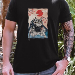 May include: Black t-shirt featuring a Japanese-inspired graphic of a cat samurai. The artwork includes a red sun, cherry blossoms, and a mountain scene. The cat is depicted in traditional samurai attire.