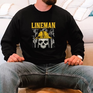 May include: Black sweatshirt with a skull wearing a yellow hard hat and the text "LINEMAN" printed on the front. The skull is surrounded by power lines and poles.