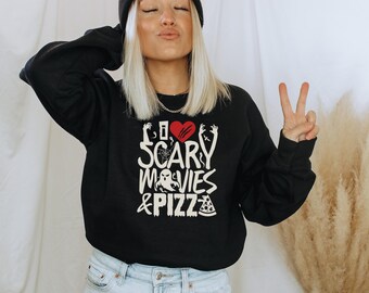 May include: A black long-sleeve sweatshirt featuring a graphic print with the text I HEART SCARY MOVIES & PIZZA. The design includes a red heart, a white ghost, a spider web, and a slice of pizza with white lettering on a black background.