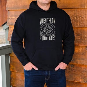 Puede incluir: Sudadera con capucha negra con un estampado gráfico blanco que dice "When the DM smiles it's already too late." El gráfico presenta un dado estilizado con el número 20 en la parte superior y 8 en la parte inferior.
