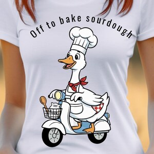 May include: A white t-shirt featuring a graphic of a white goose wearing a chef hat and red neckerchief, riding a white scooter. The goose has a basket containing a wooden spoon and a bag labeled flour. The text reads Off to bake sourdough.