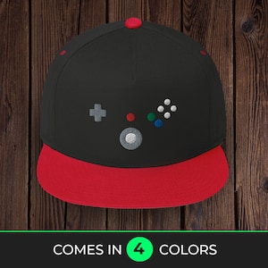 May include: Black and red baseball cap with an embroidered video game controller design. The cap features a flat brim and a red button on top. The text "COMES IN 4 COLORS" is at the bottom of the image.
