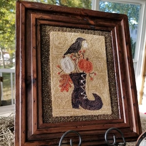 May include: A framed needlepoint artwork depicting a black crow perched on a branch with pumpkins and fall leaves in a boot. The artwork is in a dark brown wooden frame and is displayed on a stand. The tag below the artwork reads "Sew Cherished".