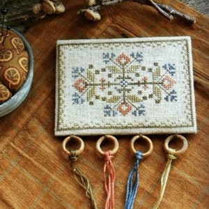 May include: A cross-stitch sampler with a geometric design in shades of brown, blue, and orange. The sampler is mounted on a wooden frame and has four tassels hanging from the bottom.