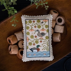 May include: A cross-stitch sampler pillow featuring a pair of ducks in a pond setting. The pillow is in a wooden bowl with spools of thread. The text "Quack Stack" and "Original Designs by Paulette Stewart" are at the bottom of the image.