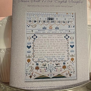 May include: A reproduction of a sampler created by Susan Scott in 1734. The sampler features a colorful alphabet, a verse from Ephesians, and the Lord's Prayer. The sampler is framed and has a white background.