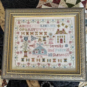 May include: Framed cross-stitch sampler with a gold-colored ornate frame. The sampler features a house, floral elements, and the text "Serenity is all She needs." A pair of decorative scissors rests below the frame.