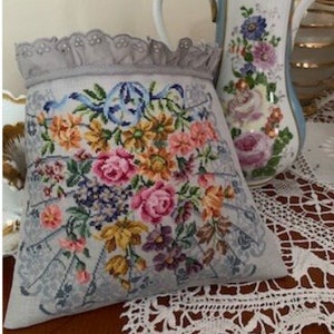 May include: A gray and white pillow with a floral embroidery design. The pillow has a lace trim and is sitting on a white crocheted doily.