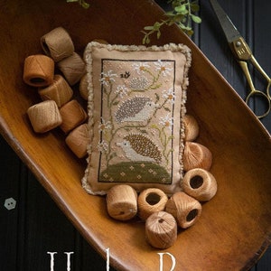 May include: A finished cross-stitch sampler titled "Hedge Row" by Plum Street Samplers. The sampler features two hedgehogs in a field of flowers. The sampler is displayed in a wooden bowl with brown embroidery floss spools.