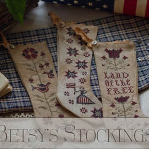 May include: Three embroidered stockings with patriotic themes. The stockings are beige with red, white, and blue embroidery. One stocking features a star, another features a bird, and the third features a flower and the text "Land of the Free".