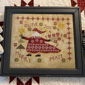 May include: A framed cross-stitch design featuring a red and white Santa Claus holding a bouquet of flowers. The text "Believe Joy Merry Peace" is stitched in the background.