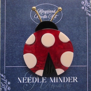 May include: A ladybug-shaped needle minder with a red body and white spots. The needle minder has a black head and a black triangle at the bottom. The text "Kingsland Needle Art" and "Needle Minder" are visible.