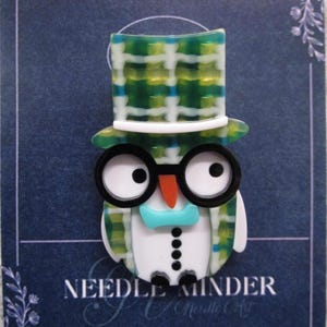 May include: A whimsical needle minder in the shape of an owl wearing a plaid top hat and glasses. The owl has a white body, a turquoise bow tie, and a small orange beak. The text "NEEDLE MINDER" is visible.