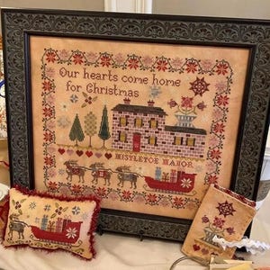 May include: A framed cross-stitch sampler with a red and white color scheme. The sampler features a house with a red door and the text "Our hearts come home for Christmas" and "Mistletoe Manor".