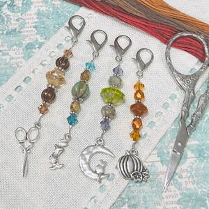 May include: Four colorful beaded stitch markers with different charms, including a crescent moon, a pumpkin, a bunny, and a pair of scissors. The stitch markers are hanging from silver rings.