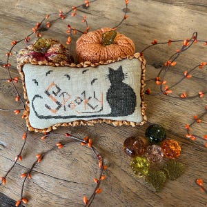 May include: A beige pillow with a black cat and the word "Spooky" embroidered on it. The pillow is surrounded by orange and brown fall decorations.