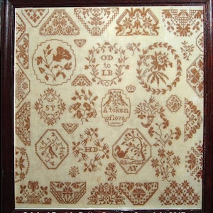 May include: A framed sampler with a white background and brown embroidery. The sampler features various designs, including flowers, birds, and geometric patterns. The text "OD to LB A token of love" is embroidered in the center.