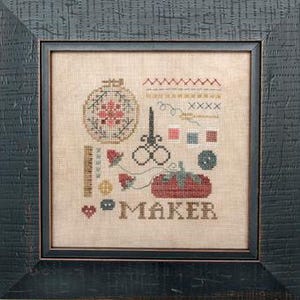 May include: Cross-stitch sampler with a black frame. The design includes a floral embroidery hoop, scissors, a tomato, and the word "MAKER." Various stitches, buttons, and a heart are also featured. The color palette includes red, pink, blue, and brown.