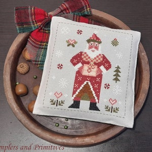 May include: A white cross-stitch sampler with a red and white Santa Claus holding a gift. The background is white with snowflakes and a green tree. The sampler is framed with a gray border.