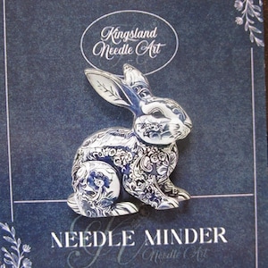 May include: A blue and white ceramic bunny needle minder with intricate floral patterns. The bunny is set against a dark blue background with the text "Kingsland Needle Art" and "Needle Minder".