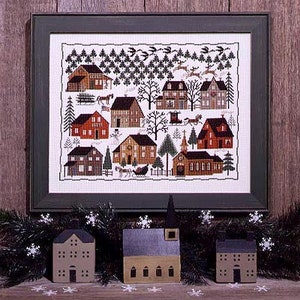 May include: A cross-stitch pattern of a winter village scene with houses, a church, and a sleigh pulled by horses. The pattern is framed in a dark green frame and is displayed on a wooden wall.
