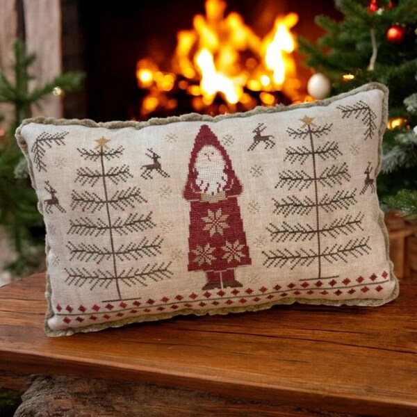 Op de afbeelding: Een decoratief kussen met een kruissteekontwerp met een rood geklede kerstman die een cadeau vasthoudt, twee groene dennenbomen en rendieren. Het kussen is gemaakt van beige linnen stof met een groene fluwelen rand.