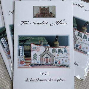 May include: Several cross-stitch pattern packets. The main packet features a schoolhouse sampler design with the text "1871 Schoolhouse Sampler." The design includes a schoolhouse, trees, and children. The packet is titled "The Scarlett House."