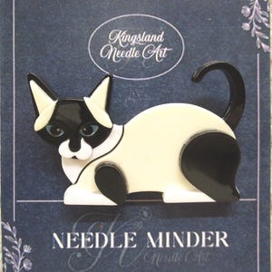 May include: A black and white cat-shaped needle minder with blue eyes, set against a dark blue background. The needle minder has the text "Kingsland Needle Art" and "Needle Minder" printed on it. The cat has a cream-colored body and black markings.