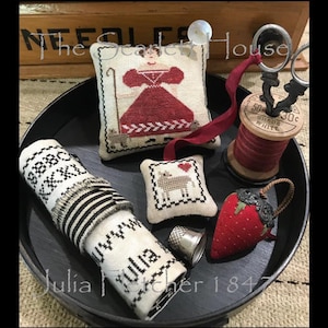 May include: A black tray with a variety of needlepoint and embroidery projects. The projects include a small pillow with a woman in a red dress, a small pillow with a sheep, a rolled-up piece of fabric with black and white stripes, a spool of red thread, and a strawberry-shaped pincushion.