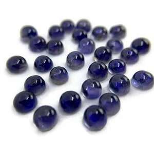 Puede incluir: Un conjunto de pequeñas gemas redondas y pulidas en tonos azul oscuro. Las gemas están dispersas sobre una superficie blanca, cada una con una superficie lisa y reflectante.
