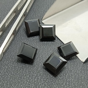 May include: Five square-cut, black gemstones are displayed on a textured surface. The gemstones are faceted, reflecting light, and are arranged near a pair of metal tools. The gems have a polished, glossy appearance.