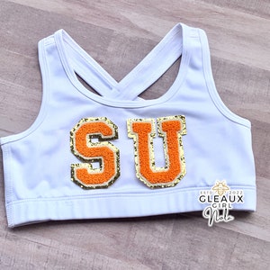 May include: White sports bra with a criss-cross back strap and the letters "SU" in orange and gold glitter on the front. The bra has the text "ESTD 2022 GLEAUX GIRL NOLA" on the bottom.