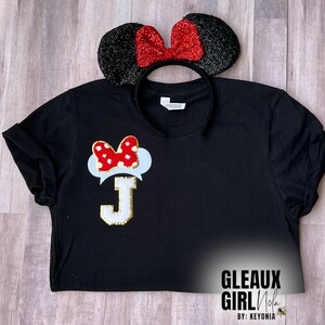 Könnte beinhalten: Schwarzes T-Shirt mit einem weißen und roten Minnie Mouse Kopf mit einem weißen Buchstaben J in der Mitte. Das Shirt wird mit einem schwarzen Stirnband mit roter Schleife und schwarzen Ohren kombiniert.