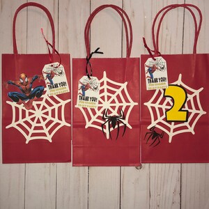 May include: Three red gift bags with white spider web designs and a Spiderman character. Each bag has a tag that says "Thank You!! For coming to my party".