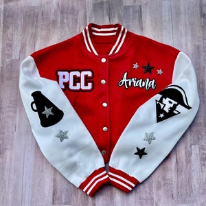 以下が含まれることがあります： PCCとArianaの文字が入った赤と白のバーシティジャケット。白い袖には黒いメガホンとマスコットのグラフィックがあり、シルバーとブラックのグリッタースターがアクセントになっています。スナップボタン式のフロントとストライプ柄のリブトリムが特徴です。