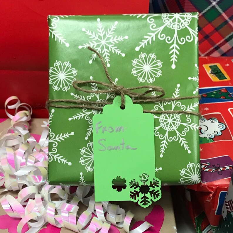 May include: A green gift box with a white snowflake pattern wrapped with twine and a green gift tag that says "From Santa".