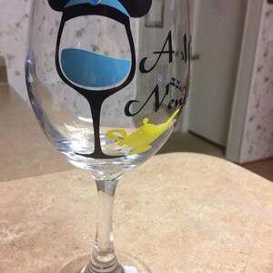 May include: A clear wine glass with a black silhouette of a wine glass with blue liquid inside. The glass has a black Mickey Mouse ears design with a blue hat and a yellow genie lamp on the side. The text "A Whole New" is partially visible on the glass.