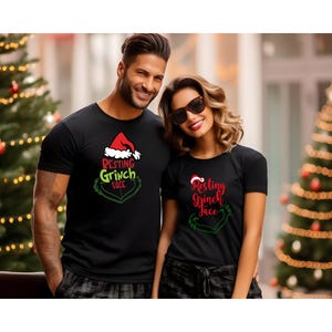 May include: Two people wearing black t-shirts with a Christmas-themed graphic. The shirts feature the text "Resting Grinch Face" in red and green, along with a Santa hat and Grinch face design. Christmas trees are in the background.