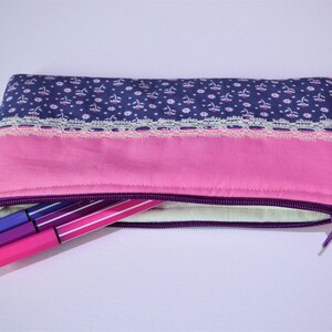 May include: A purple and pink fabric pencil case with a zipper closure. The case has a floral pattern and a lace trim. Four purple and pink pens are sticking out of the zipper opening.