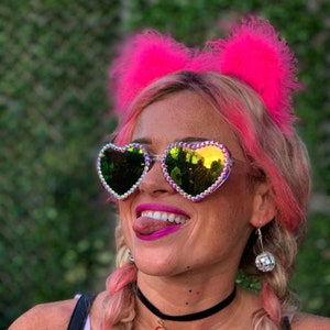 May include: A woman with pink hair is wearing heart-shaped sunglasses with iridescent rhinestones. She is also wearing a pink feather headband and a black choker necklace with a rainbow charm.