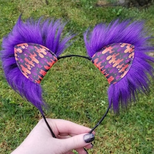May include: A pair of cat ears with purple faux fur and patterned ear inserts. The inserts feature a black background with orange and red flame-like designs. The ears are attached to a black headband.