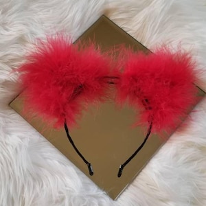 May include: Red feather headband with a black headband. The headband is on a white fluffy surface.