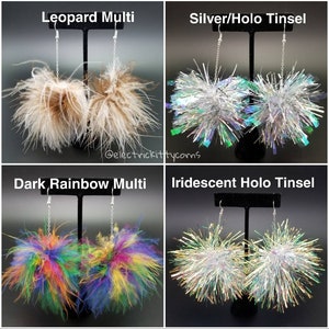 May include: Four pairs of dangle earrings with pom-pom accents. The earrings are displayed on a black stand. The pom-poms are in the following colors: leopard print, silver with iridescent holo tinsel, dark rainbow multi, and iridescent holo tinsel.